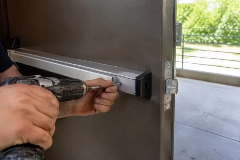 Hands using a power driver at the end of a horizontal push bar on an open metal door. — Panic & Exit Hardware, Toronto